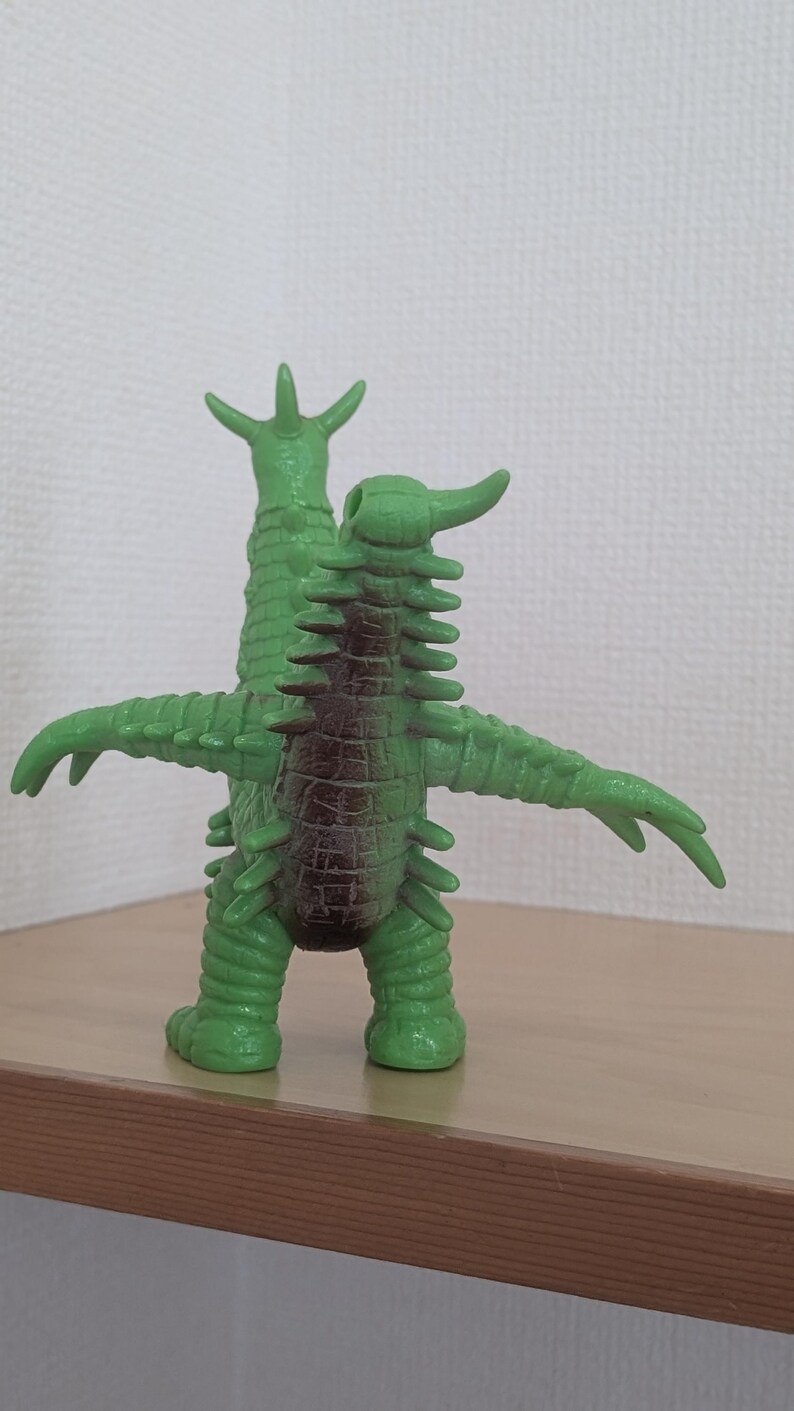 May include: A green monster figurine with two heads, multiple spikes along its back and arms, and a dark brown patch on its torso. The creature stands upright with its arms extended outward, featuring a textured, reptilian-like skin surface.