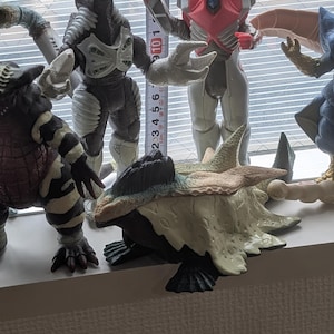 Op de afbeelding: Een verzameling van vijf plastic monsteractiefiguren. Een meetlint is verticaal achter de figuren geplaatst, waaruit blijkt dat het langste figuur ongeveer 21,5 cm hoog is. De figuren hebben verschillende kleuren, waaronder zwart, wit, roze en beige met gedetailleerde texturen.