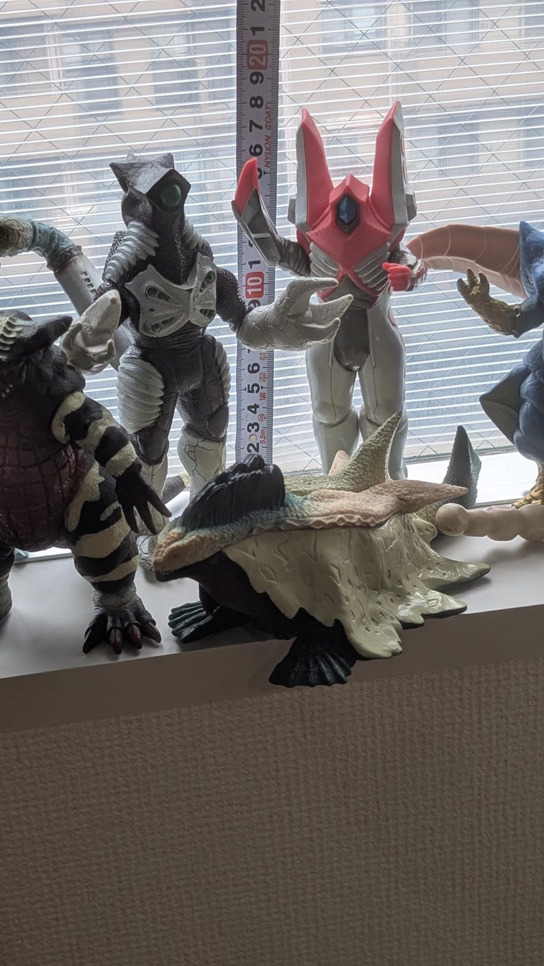 Op de afbeelding: Een verzameling van vijf plastic monsteractiefiguren. Een meetlint is verticaal achter de figuren geplaatst, waaruit blijkt dat het langste figuur ongeveer 21,5 cm hoog is. De figuren hebben verschillende kleuren, waaronder zwart, wit, roze en beige met gedetailleerde texturen.