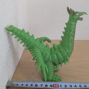 May include: A green, reptilian-like monster figurine with a long neck, spiked tail, and textured skin. The figure is positioned next to a metal measuring tape, which indicates a length of approximately 6.5 inches from tail to chest.