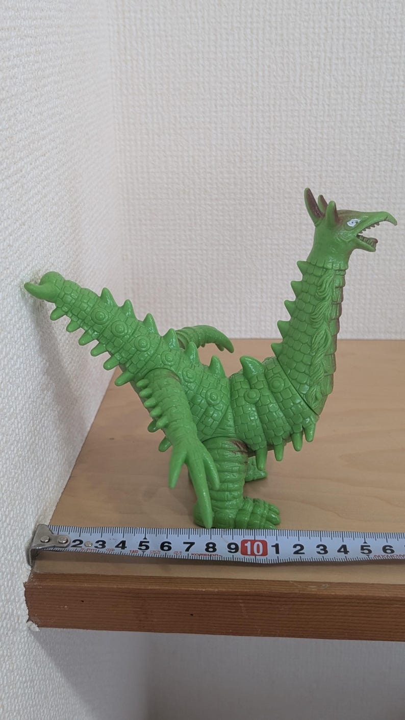 May include: A green, reptilian-like monster figurine with a long neck, spiked tail, and textured skin. The figure is positioned next to a metal measuring tape, which indicates a length of approximately 6.5 inches from tail to chest.