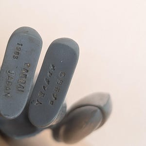 May include: Close-up of the feet of a gray action figure. The soles of the feet are visible, with the text "1983 BANDAI JAPAN" and Japanese characters. The figure is on a light background.