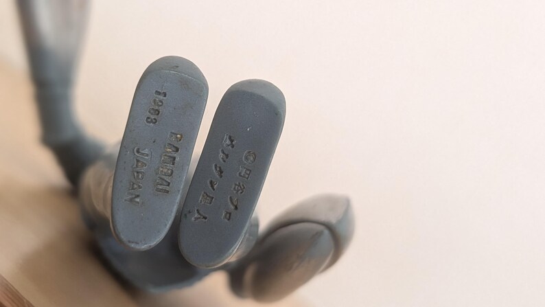 May include: Close-up of the feet of a gray action figure. The soles of the feet are visible, with the text "1983 BANDAI JAPAN" and Japanese characters. The figure is on a light background.