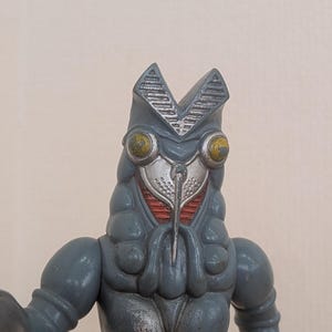 May include: A gray, stylized monster figure with a detailed head, large yellow eyes, and a silver faceplate. The figure has a textured body and arms, and is a collectible toy.