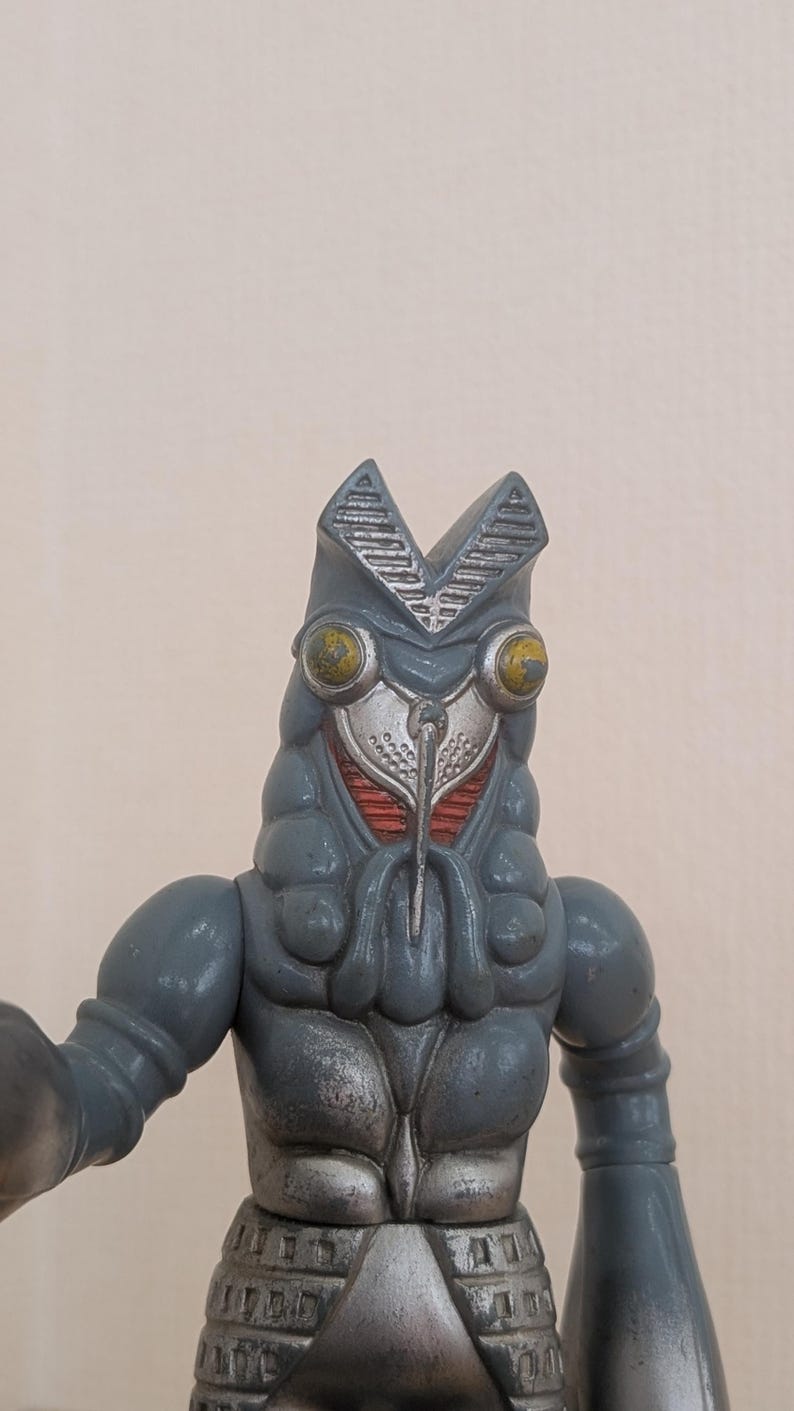 May include: A gray, stylized monster figure with a detailed head, large yellow eyes, and a silver faceplate. The figure has a textured body and arms, and is a collectible toy.
