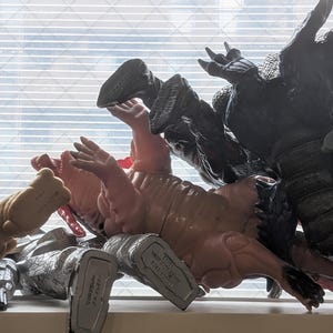 Op de afbeelding: Een verzameling van diverse plastic monsteractiefiguren in verschillende kleuren, waaronder beige, roze, zwart en zilver, geplaatst in een rommelige hoop met hun voeten en ledematen omhoog gericht naar een raam met horizontale jaloezie&euml;n.