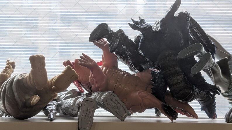 Op de afbeelding: Een verzameling van diverse plastic monsteractiefiguren in verschillende kleuren, waaronder beige, roze, zwart en zilver, geplaatst in een rommelige hoop met hun voeten en ledematen omhoog gericht naar een raam met horizontale jaloezie&euml;n.