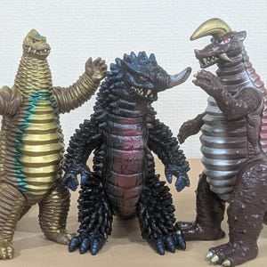May include: Three kaiju monster action figures standing side by side. The left figure is gold with blue accents, the center figure is black with a single horn, and the right figure is brown with a silver chest and a curved horn on its head.