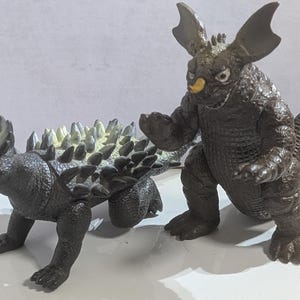 May include: Two dark brown monster figurines. One is a quadrupedal creature with a textured back covered in light-colored, pointed spikes. The other is a bipedal creature with large, wing-like ears, a small yellow horn on its snout, and a long tail.
