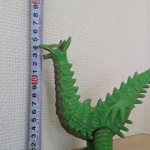 May include: A green monster figurine with a long neck, textured scales, and sharp spikes along its back and tail. A vertical measuring tape placed next to the figure indicates a height of approximately 23.5 centimeters from the base to the head.