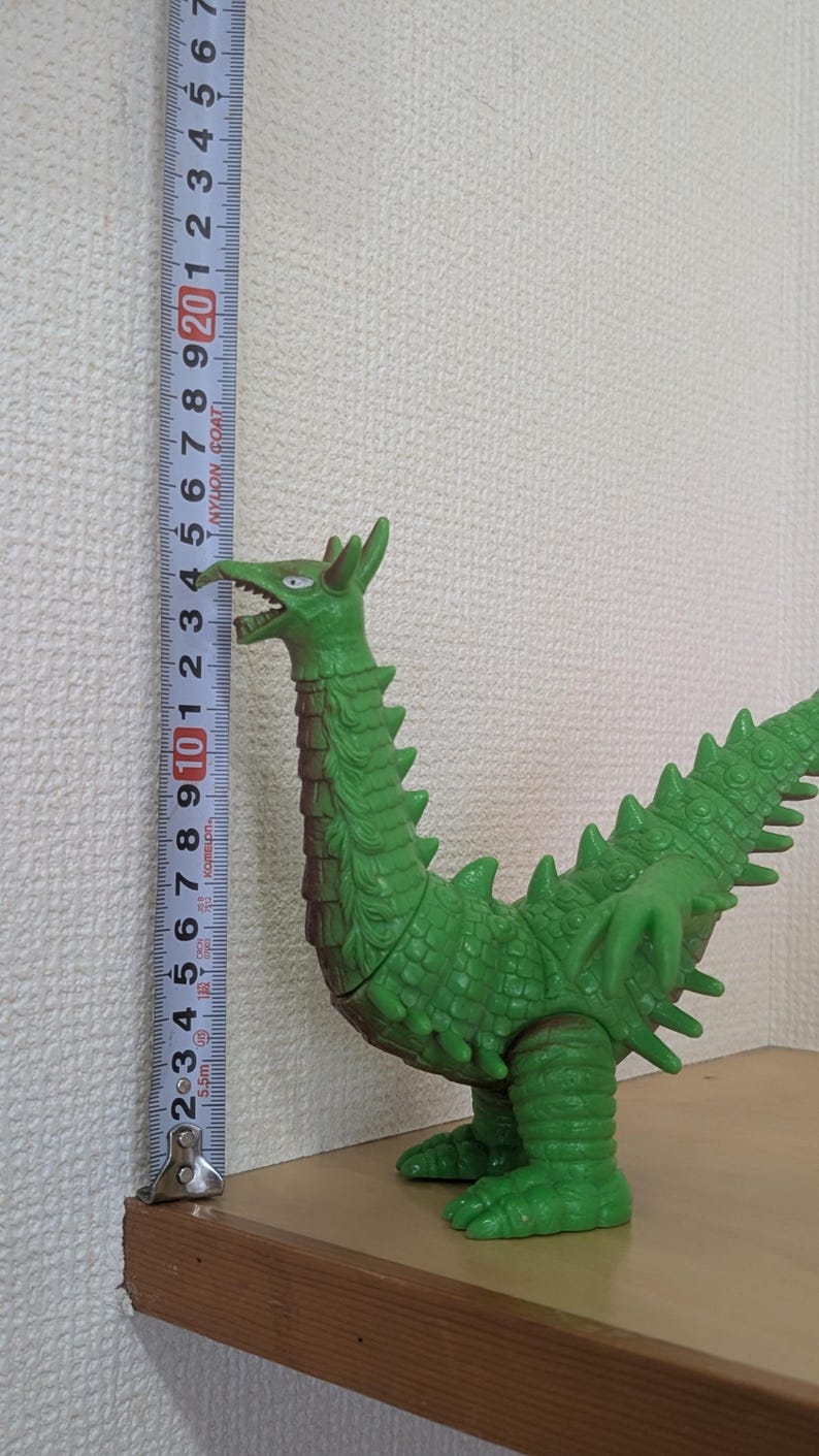 May include: A green monster figurine with a long neck, textured scales, and sharp spikes along its back and tail. A vertical measuring tape placed next to the figure indicates a height of approximately 23.5 centimeters from the base to the head.