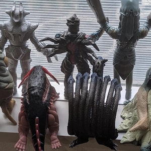 Op de afbeelding: Een verzameling van negen diverse monsteractiefiguren die in twee rijen staan. De figuren hebben verschillende texturen, kleuren waaronder zilver, zwart, rood en beige, en duidelijke anatomische vormen, waaronder enkele met meerdere ledematen.