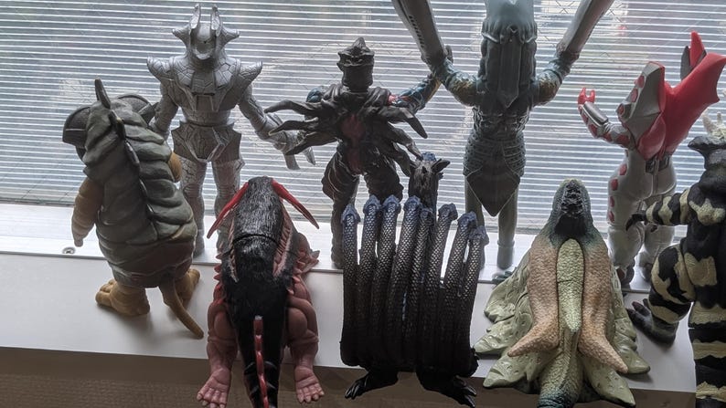 Op de afbeelding: Een verzameling van negen diverse monsteractiefiguren die in twee rijen staan. De figuren hebben verschillende texturen, kleuren waaronder zilver, zwart, rood en beige, en duidelijke anatomische vormen, waaronder enkele met meerdere ledematen.