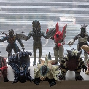 Op de afbeelding: Een verzameling van twaalf diverse monster-actiefiguren in verschillende kleuren en vormen, waaronder humano&iuml;de en wezenachtige ontwerpen. De figuren zijn in twee rijen gerangschikt en vertonen gedetailleerde texturen, hoorns, klauwen en vleugels.