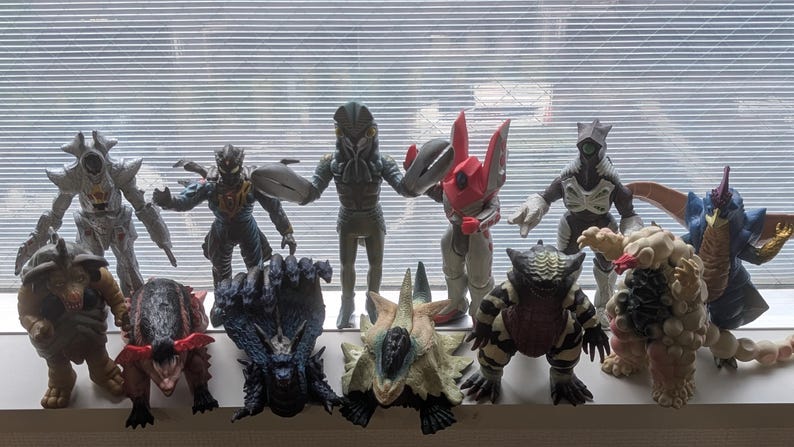 Op de afbeelding: Een verzameling van twaalf diverse monster-actiefiguren in verschillende kleuren en vormen, waaronder humano&iuml;de en wezenachtige ontwerpen. De figuren zijn in twee rijen gerangschikt en vertonen gedetailleerde texturen, hoorns, klauwen en vleugels.
