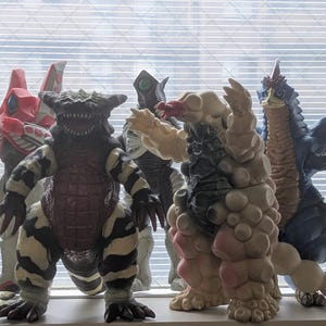 Op de afbeelding: Een verzameling van zes kleurrijke monsteractiefiguren die op een rij staan. De figuren hebben verschillende ontwerpen, waaronder een grijs gevleugeld wezen, een rood-witte robotachtige figuur, een zwart-wit gestreept beest en een beige monster.