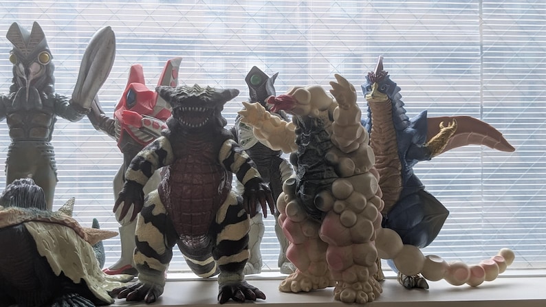 Op de afbeelding: Een verzameling van zes kleurrijke monsteractiefiguren die op een rij staan. De figuren hebben verschillende ontwerpen, waaronder een grijs gevleugeld wezen, een rood-witte robotachtige figuur, een zwart-wit gestreept beest en een beige monster.