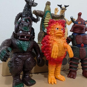 May include: A collection of five colorful monster action figures. The group includes a dark brown creature with green accents, a bright orange and red textured figure, a dark red armored character, a tall brown reptilian figure, and a black horned beast.