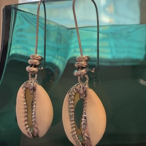 Puede incluir: Un par de pendientes colgantes con conchas de cauri blancas naturales con adornos de metal plateado. Los pendientes tienen un diseño en espiral encima de la concha y un gancho largo y delgado. El fondo es de cristal color turquesa.