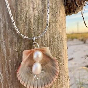 May include: A silver necklace featuring a seashell pendant with two white pearls. The seashell is a light pink and cream color, with a textured surface. The necklace is displayed against a wooden post with a beach background.