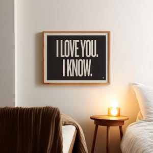 May include: A framed print with the text "I LOVE YOU. I KNOW." in white on a black background. The print is in a light wood frame and hangs on a white wall. The scene includes a bed and a small wooden side table with a lamp.