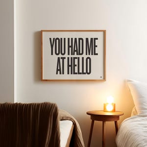 Peut inclure: Impression encadrée avec le texte "YOU HAD ME AT HELLO" en lettres noires grasses. L'impression est dans un cadre en bois clair et est accrochée à un mur blanc. Une petite table d'appoint en bois avec une lampe se trouve en dessous.
