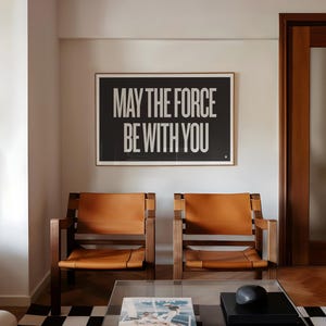 Puede incluir: Un póster enmarcado en blanco y negro con el texto "MAY THE FORCE BE WITH YOU" cuelga sobre dos sillas de madera con asientos de cuero marrón. Las sillas están en un salón.