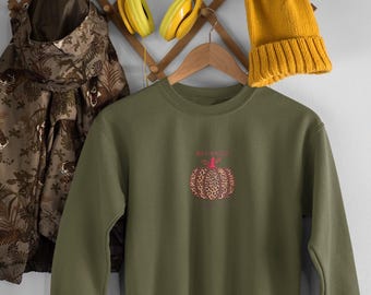 Puede incluir: Sudadera verde oliva con un diseño de calabaza con estampado de leopardo y la palabra "BLESSED" encima. Un gorro y unos auriculares amarillos cuelgan de un perchero de pared de madera. Una chaqueta estampada cuelga a la izquierda.