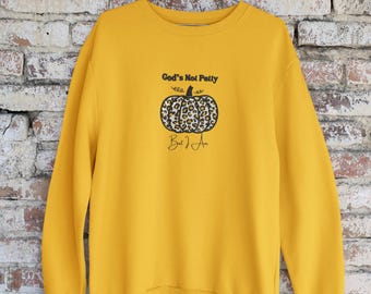 Puede incluir: Sudadera amarilla mostaza con un diseño de calabaza. La calabaza tiene un estampado de leopardo y el texto "God's Not Petty, But I Am" en negro. La sudadera está colgada en una percha de metal contra una pared de ladrillos.
