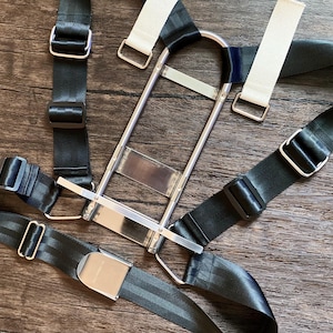 Puede incluir: Un arnés de cinturón de seguridad de metal plateado y negro con correas blancas y negras. El arnés tiene hebillas de metal y una placa rectangular de metal. Las correas están hechas de un material tejido duradero.