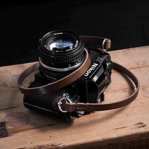 May include: A black Nikon camera with a lens and a brown leather strap. The camera is on a wooden surface. The strap is looped around the camera body.
