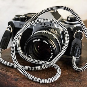 Puede incluir: Una cámara Nikon plateada con una lente negra y una correa a cuadros en blanco y negro. La lente dice "NIKKOR 50mm 1:1.4". La cámara está sobre una superficie de madera.