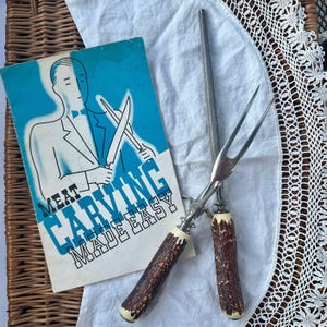 May include: Vintage meat carving set with a sharpening steel and a booklet. The carving fork and knife handles have a brown, textured design with cream-colored accents. The booklet has a blue background with the text "MEAT CARVING MADE EASY."