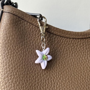 May include: A lavender flower charm with a silver-colored clasp and chain is attached to a brown leather bag. The flower has a green and brown center. The bag has a textured surface.