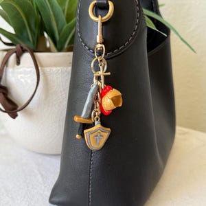 Op de afbeelding: Een goudkleurige metalen sleutelhanger bevestigd aan een zwarte handtas. De sleutelhanger bevat een kleine kruisbedel, een miniatuur grijs zwaard met een bruin gevest, een rood-gele helmbedel en een beige schildbedel met een grijs kruis.