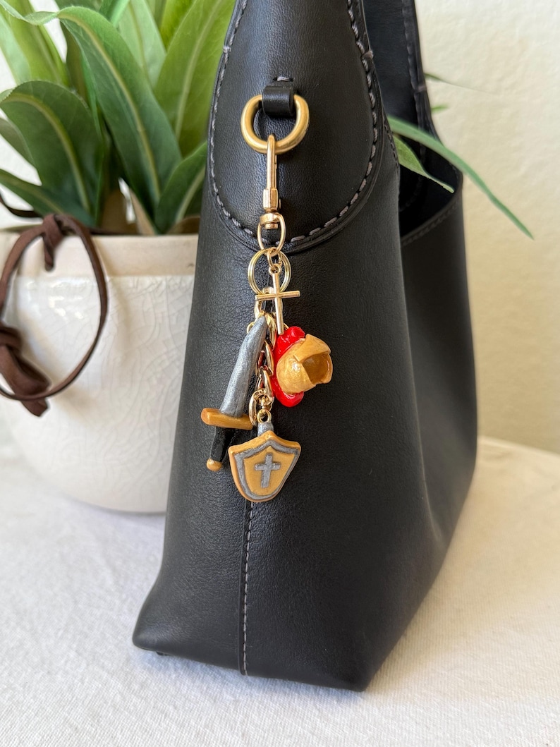 Op de afbeelding: Een goudkleurige metalen sleutelhanger bevestigd aan een zwarte handtas. De sleutelhanger bevat een kleine kruisbedel, een miniatuur grijs zwaard met een bruin gevest, een rood-gele helmbedel en een beige schildbedel met een grijs kruis.