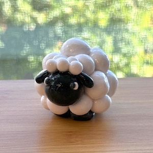 Może przedstawiać: Ręcznie wykonana ceramiczna figurka owcy. Owca ma czarną twarz i nogi, z puszystym korpusem wykonanym z białych, zaokrąglonych kształtów. Figurka jest przedmiotem dekoracyjnym, prawdopodobnie do ekspozycji.