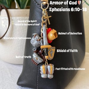 Op de afbeelding: Een goudkleurige metalen sleutelhanger met miniatuurbedels die de wapenrusting van God voorstellen, waaronder een zwaard, helm, schild, borstplaat, riem en laarzen. De tekst Armor of God Ephesians 6:10-18 staat bij de bedels afgebeeld.