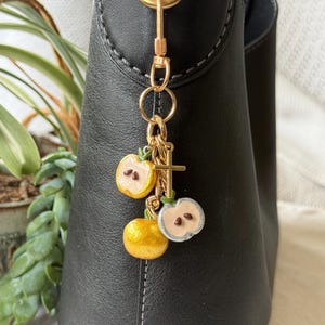 May include: A gold-toned charm with three apple-shaped pendants and a small cross, attached to a black leather bag. The apple charms are yellow and white with green stems. The bag has a gold-colored clasp.