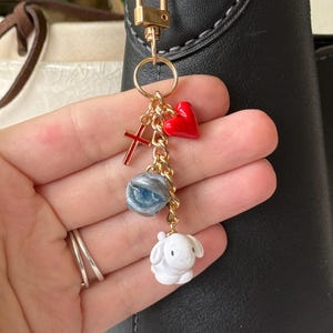 May include: A gold-colored keychain with a lobster clasp, holding charms of a red heart, a red cross, a blue and silver eye, and a white bunny. The keychain is attached to a black bag.