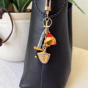 Op de afbeelding: Een goudkleurige metalen sleutelhanger bevestigd aan een zwarte leren tas. De sleutelhanger heeft drie miniatuurbedels: een zilveren en gouden zwaard, een gele en rode helm, en een geel schild met een zilveren kruis in het midden.