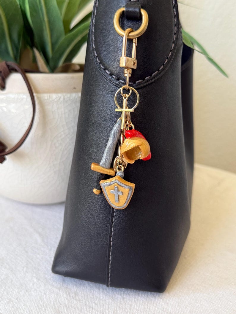 Op de afbeelding: Een goudkleurige metalen sleutelhanger bevestigd aan een zwarte leren tas. De sleutelhanger heeft drie miniatuurbedels: een zilveren en gouden zwaard, een gele en rode helm, en een geel schild met een zilveren kruis in het midden.