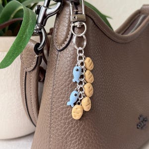 May include: A brown leather handbag with a silver-tone metal clasp and a decorative keychain. The keychain features two light blue fish charms and several tan-coloured, rounded charms. The bag has a pebbled texture.