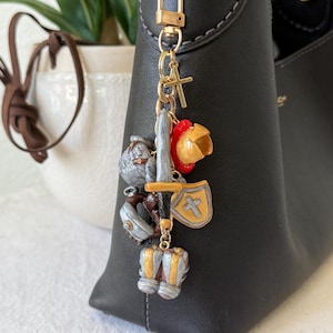 Op de afbeelding: Een goudkleurige metalen sleutelhanger met diverse miniatuurbedels, waaronder een zilverkleurig zwaard, een goudkleurig schild met een kruis, een rood-gouden helm, een zilverkleurige helm en zilverkleurige laarzen, bevestigd aan een gouden karabijnhaak.