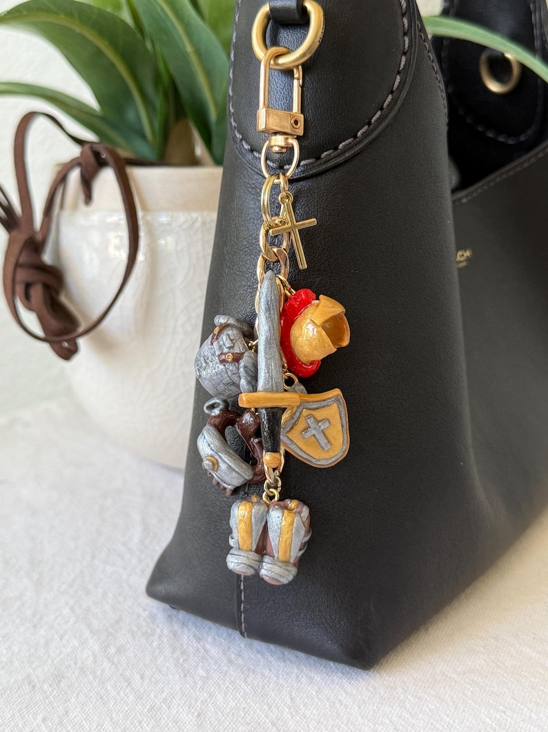 Op de afbeelding: Een goudkleurige metalen sleutelhanger met diverse miniatuurbedels, waaronder een zilverkleurig zwaard, een goudkleurig schild met een kruis, een rood-gouden helm, een zilverkleurige helm en zilverkleurige laarzen, bevestigd aan een gouden karabijnhaak.