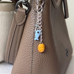Pode incluir: Uma mala de couro castanho com uma corrente prateada e um pequeno berloque. O berloque apresenta um peixe azul e uma cenoura laranja. A mala tem uma alça de ombro e um fecho de correr.