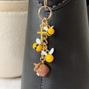 May include: A gold-colored charm with a ring, a cross, and three bee-shaped pendants. The bees are yellow and black with white wings. A brown honey pot charm hangs below the bees. The charm is attached to a black object.