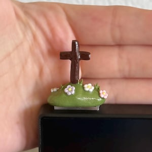 Puede incluir: Una pequeña cruz marrón se alza sobre un pequeño montículo verde adornado con diminutas flores blancas y rosas. El artículo religioso artesanal se encuentra sobre un fondo neutro.