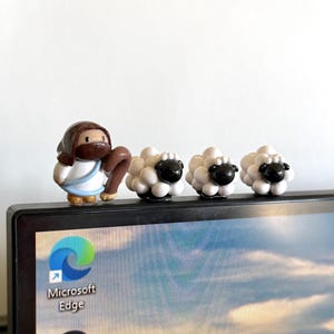 May include: A figurine of a person in a white and blue robe with a brown staff, alongside three white sheep figurines with black faces. The figurines are placed on top of a computer monitor.