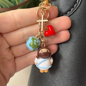 May include: A gold-toned keychain with religious charms. The charms include a cross, a red heart, a blue and green globe, and a small figurine of Jesus wearing a mask. The keychain is attached to a black leather bag.